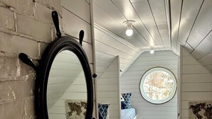 A narrow hallway with a brick wall on one side and a white paneled wall on the other, leading to a bedroom with a bed and a round window.