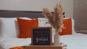 Ein ordentlich gemachtes Bett mit einer orangefarbenen Tagesdecke und Kissen, ein gerahmtes Zitat und eine Vase mit Trockenblumen auf einem Nachttisch.