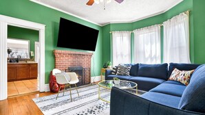 A living room with a blue sofa, a fireplace, a TV mounted on the wall, and a kitchen visible through an open door.