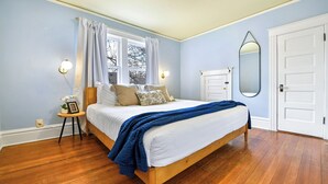 A bedroom with a wooden bed, white bedding, a blue throw blanket, a small bedside table with a lamp, and a window with curtains.