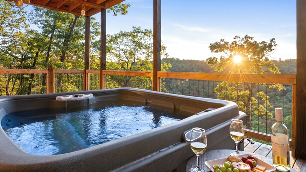 A hot tub with a wooden deck, surrounded by trees, and a table with a bottle of wine and a plate of fruit.