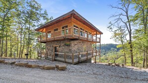 A two-story wooden cabin with a balcony, surrounded by trees and set on a gravel area.