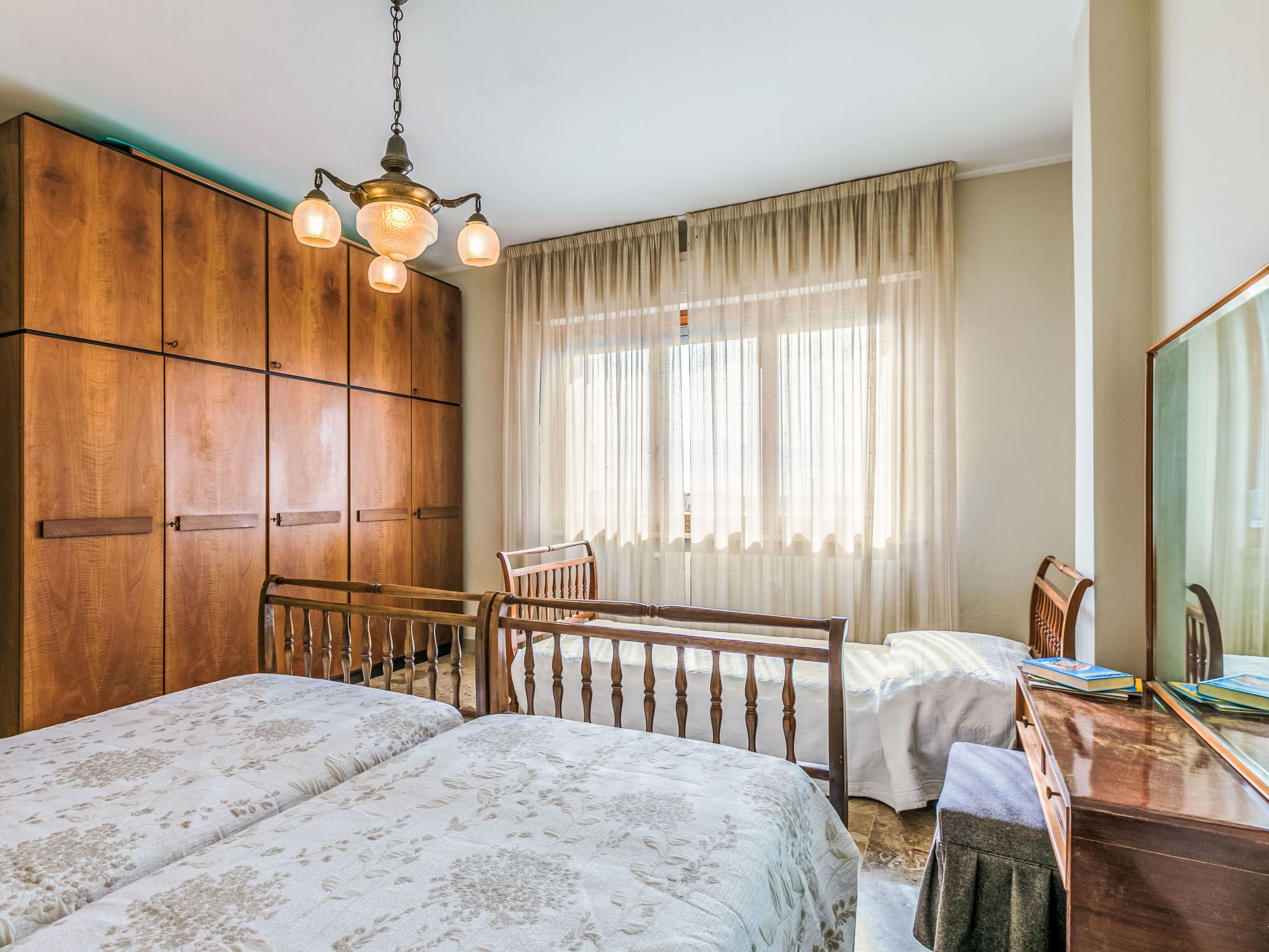 Une chambre avec deux lits, une armoire en bois, un bureau avec des livres et un grand miroir.