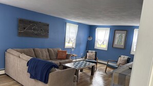 A living room with a beige sofa, a coffee table, and a blue armchair. There are two windows with sheer curtains, a floor lamp, and a map on the wall. - Charming 4-bedroom house just a Short walk to Scarborough Beach (Narragansett)