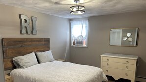 A bedroom with a wooden headboard, a white bedspread, a bedside table, a dresser, and a mirror on the wall. - Charming 4-bedroom house just a Short walk to Scarborough Beach (Narragansett)