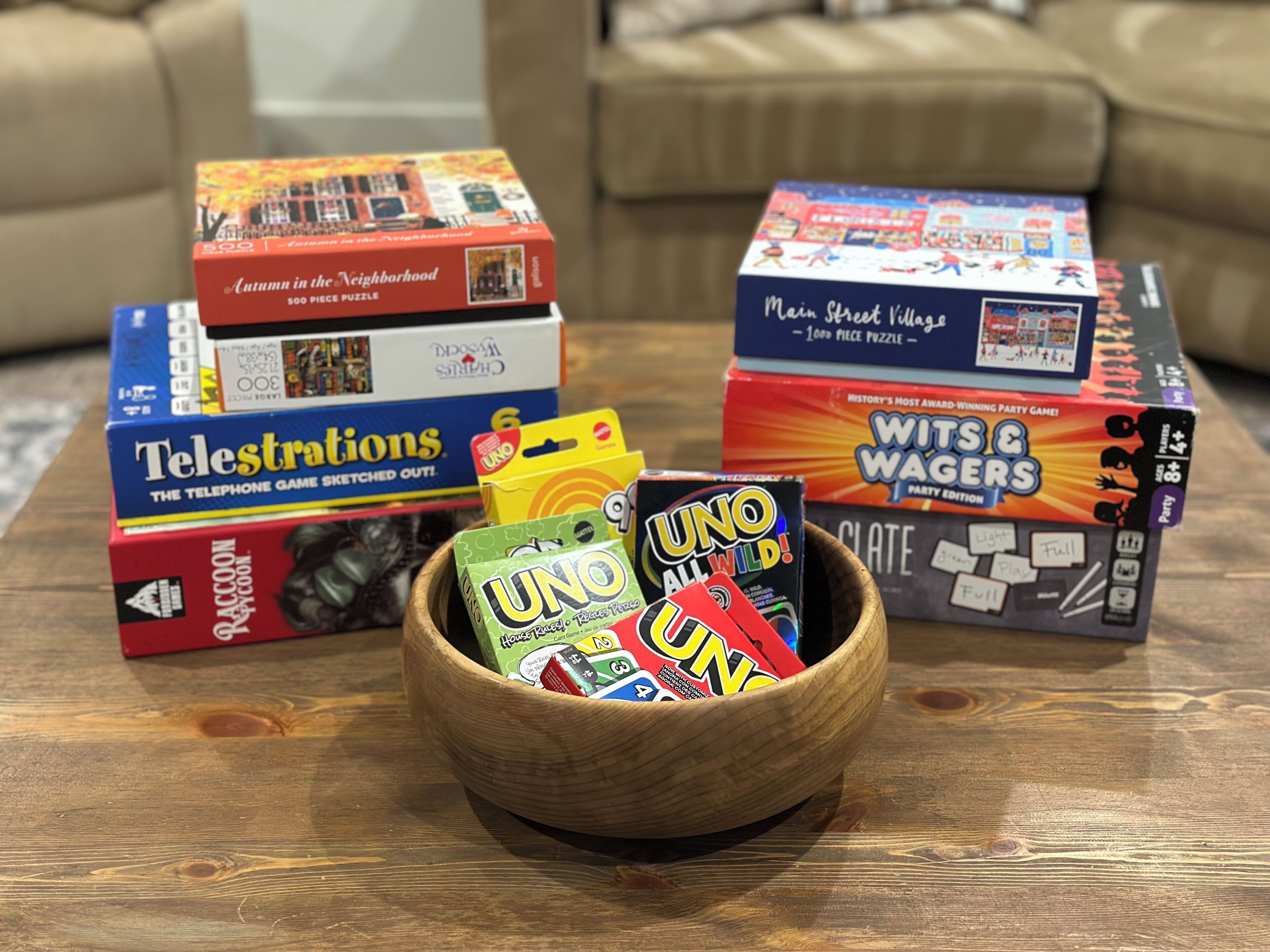 Una mesa de madera con varios juegos de mesa y rompecabezas, incluyendo 'Autumn in the Neighborhood', 'Telestrations', 'Wits & Waggers' y 'UNO'.
