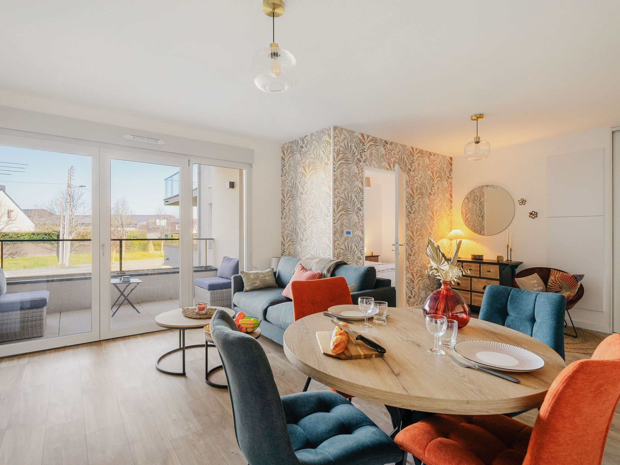 A modern living room with a large dining table, comfortable seating, and a view through a glass door to the outside.