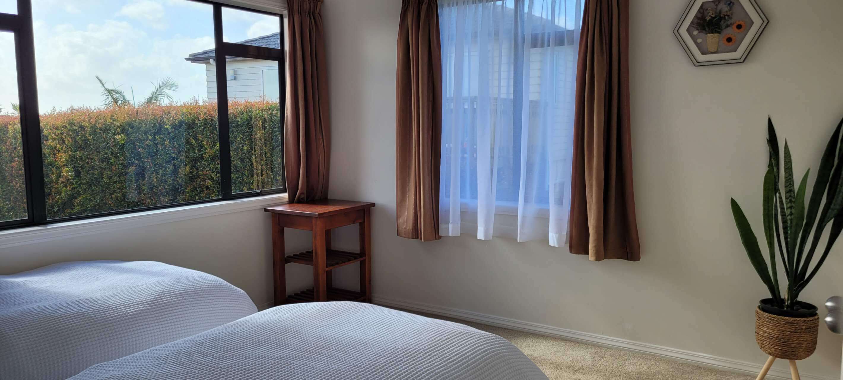Quarto com duas camas, um banquinho de madeira, uma planta em vaso e uma janela com cortinas.