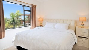 A large bed with white linens, two bedside tables with lamps, and a view of palm trees through a large window. - Elevated Family Sized Guest-house with Beautiful Views (Auckland)