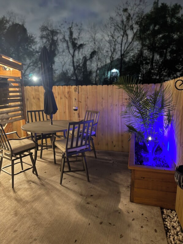An outdoor patio with a wooden table and chairs, a wooden fence, and a small planter with blue lighting.