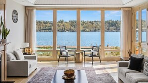 A spacious living room with a large window overlooking a lake, furnished with a sofa, chairs, and a coffee table. - Stunning Waterfront Mansion w Pool n Hottub (Mercer Island)