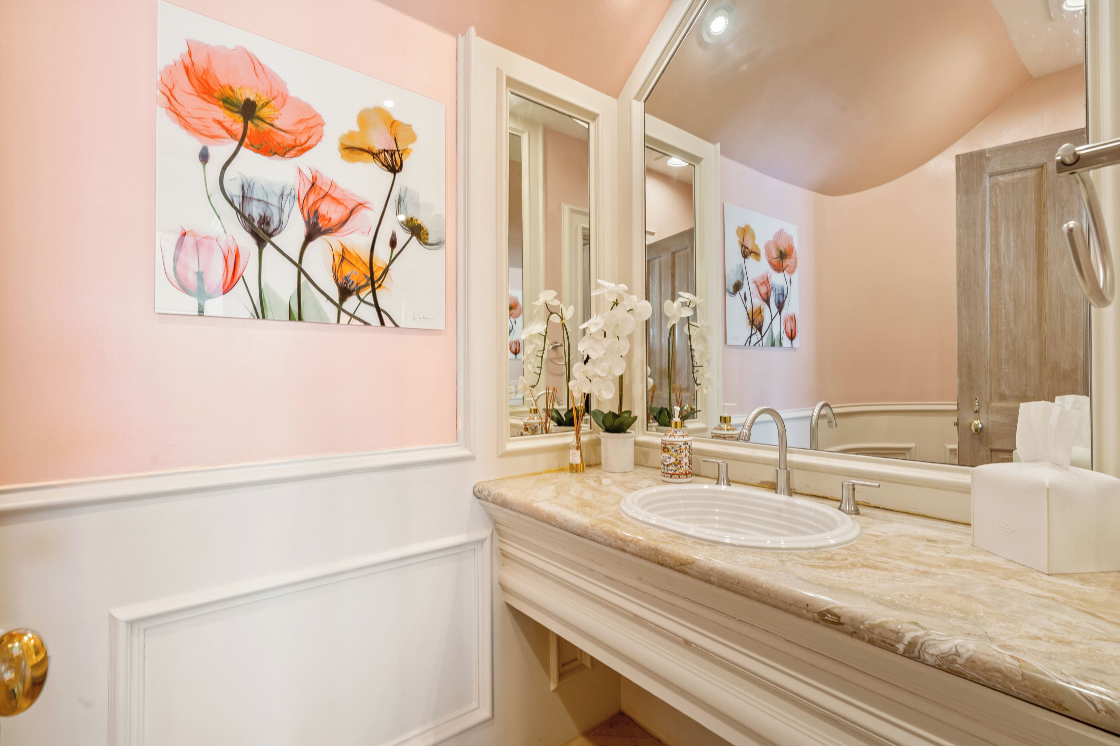 A bathroom with a white vanity, a sink, and a mirror. There are floral paintings on the wall and a vase with white flowers.