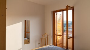 Une chambre à coucher comprenant un lit, une armoire, un miroir et une fenêtre donnant sur un immeuble et des arbres.
