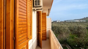 Un balcon doté de volets en bois, d’un appareil de climatisation et offrant une vue sur un paysage verdoyant, avec des bâtiments visibles au loin.