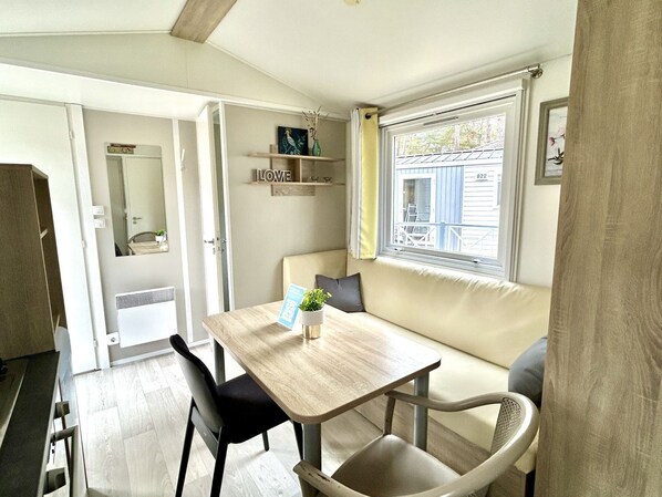 A compact living space with a dining table, chairs, a sofa, a window with a view, and a small shelf with decorative items.