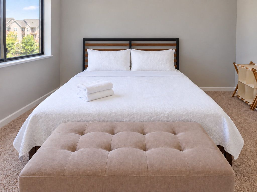 A neatly made bed with a headboard, a window with a view of buildings, and a wooden chair.