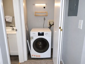 A laundry room with a front-loading washing machine, a wooden shelf, and a wall-mounted light fixture. - Recreation Central ILM (Wilmington)