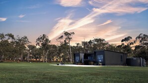 A modern, dark-colored cabin with a large window, surrounded by a grassy field and trees, under a clear sky. - Warrawindi Escapes (Penola)