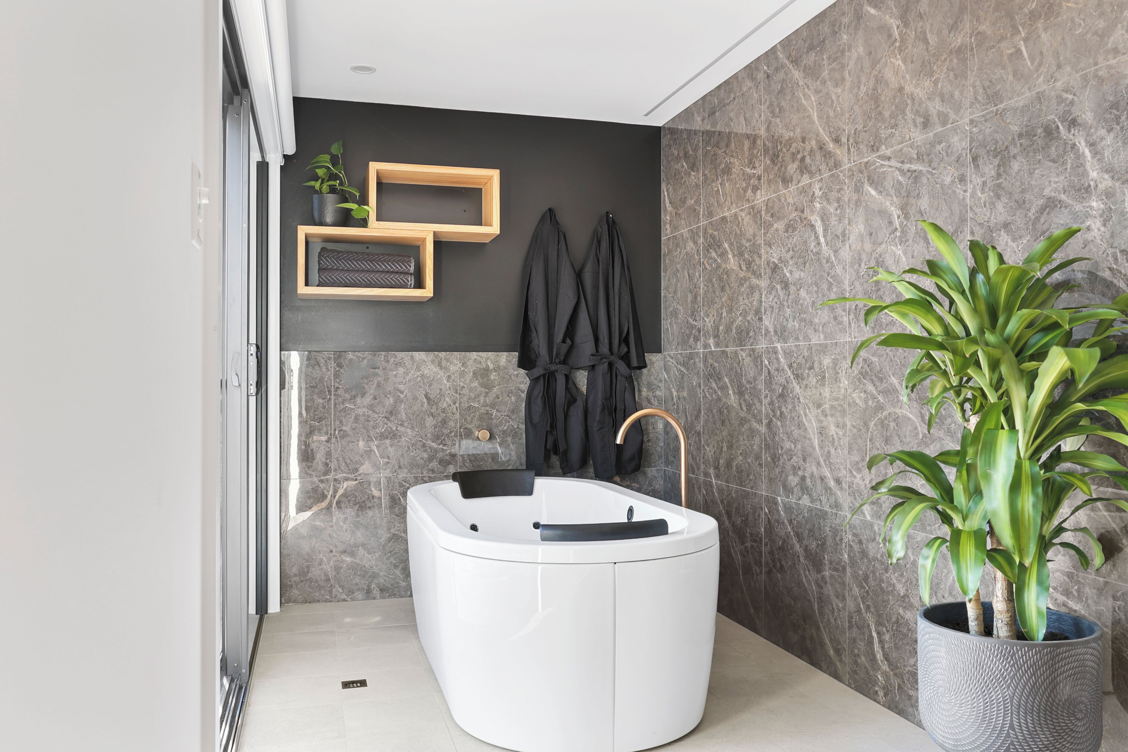 A modern bathroom with a freestanding bathtub, a potted plant, and a wooden shelf with a plant and a framed picture.