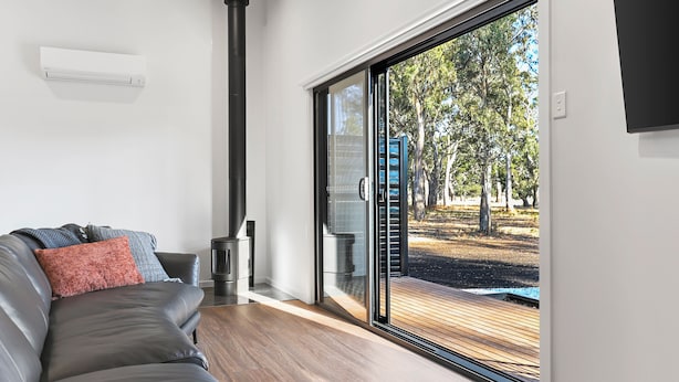 A modern living room with a leather sofa, a fireplace, and a large sliding glass door leading to an outdoor deck. - Warrawindi Escapes (Penola)