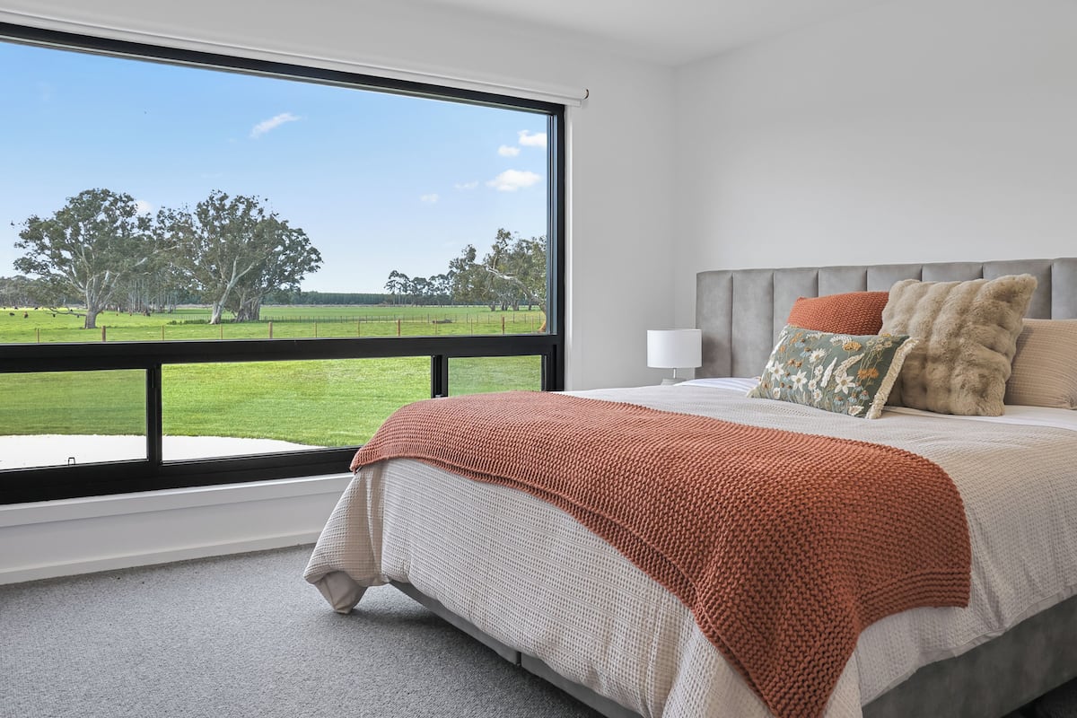 A bedroom with a large window, a bed with a grey headboard, and a view of a green field.