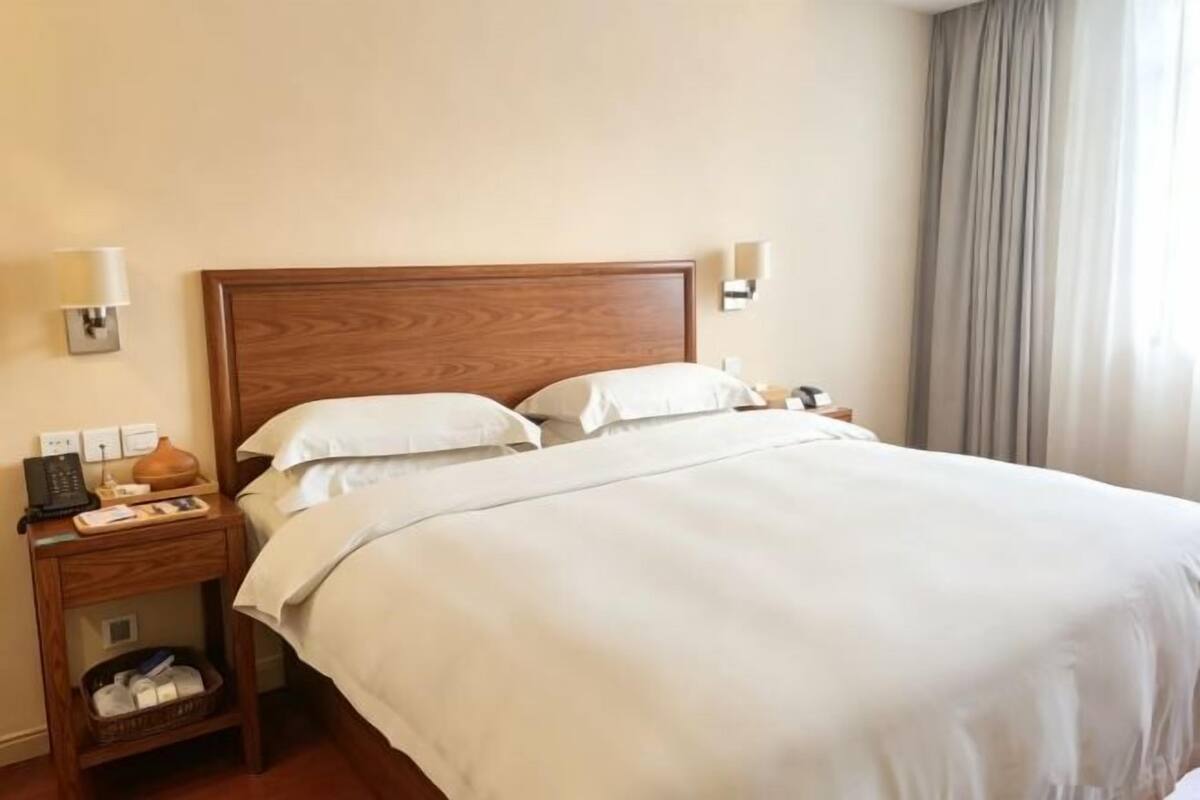 A hotel room with a wooden headboard, two white pillows, a bedside table with a lamp, and a window with curtains.