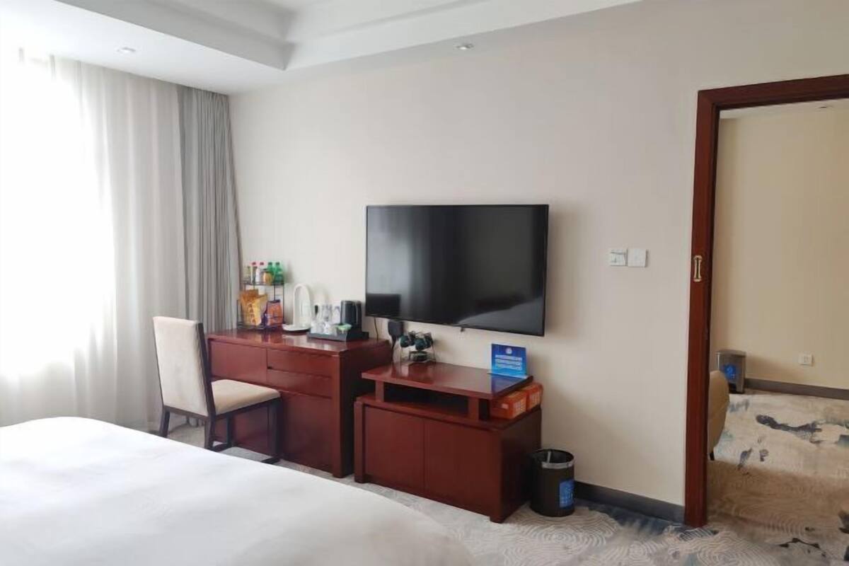 A hotel room with a bed, a desk with a flat-screen TV, a chair, and a small trash bin.