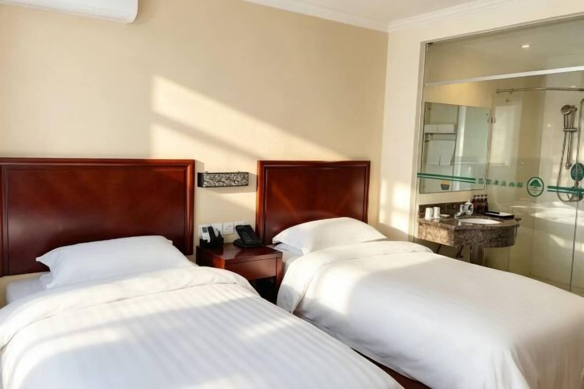 A hotel room with two beds, a bedside table with a phone, a mirror, and a bathroom visible through a glass partition.