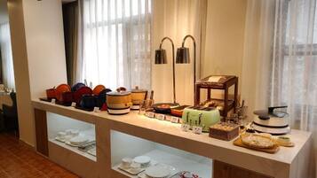 A buffet setup with various food items and containers, a lamp, and a window with curtains.