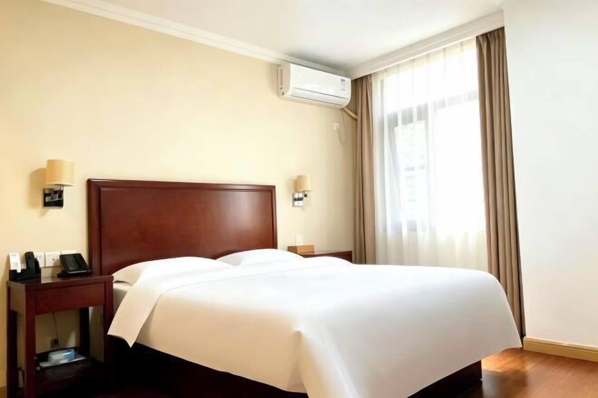 A hotel room with a large bed, bedside tables with lamps, an air conditioning unit, and a window with curtains.