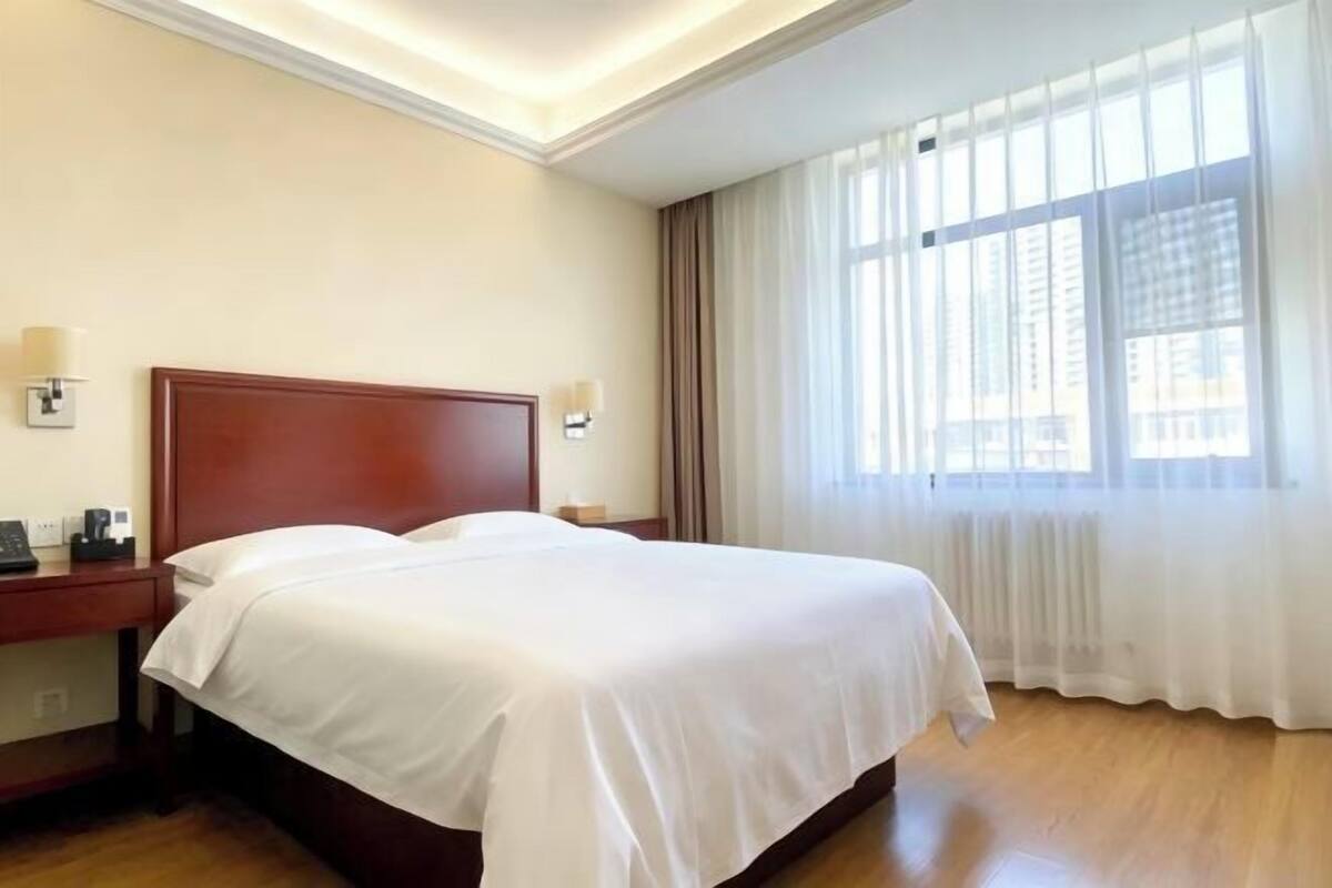A hotel room with a wooden bed, a nightstand, a lamp, and a large window with curtains.