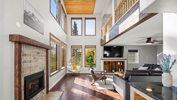 A spacious living room with a fireplace, large windows, a high ceiling, and a modern kitchen area.