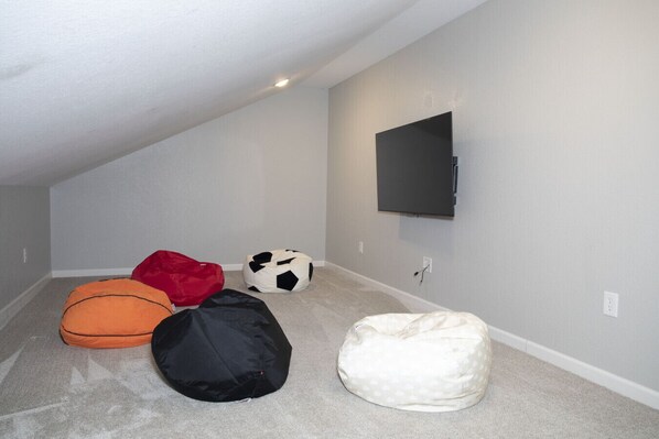 Une pièce avec un plafond en pente, un téléviseur à écran plat fixé au mur et quatre fauteuils en forme de sac de haricots de différentes couleurs.