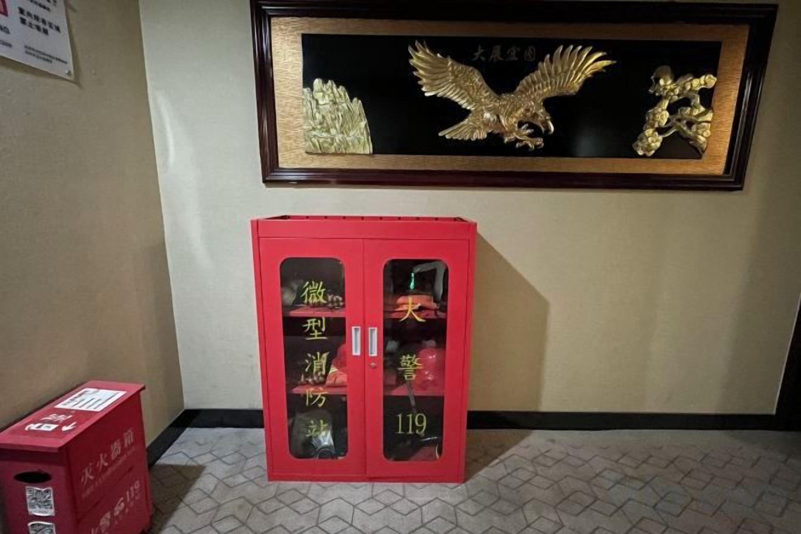 Una obra de arte enmarcada con una águila en la pared, un armario rojo con puertas de cristal y una caja roja en el suelo.