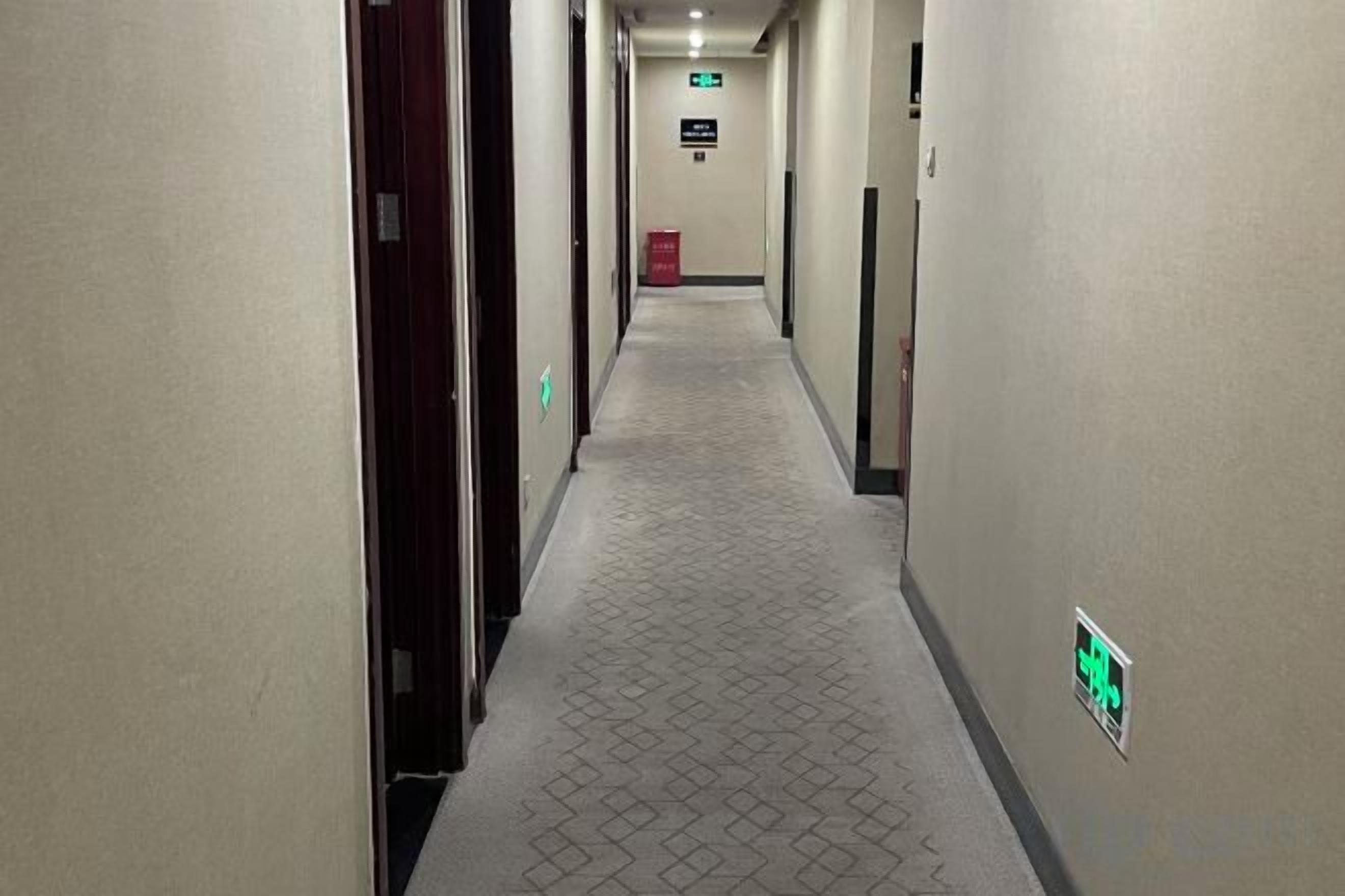 Un pasillo de hotel con puertas a ambos lados, una alfombra estampada y una señalización de salida de emergencia verde.