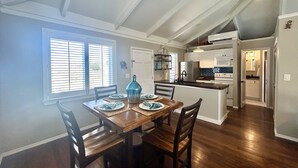 A dining area with a wooden table and chairs, a kitchen with white cabinets and appliances, and a view of the outdoors through a window with blinds. - NEW! Saltwater Serenity - Poolside & Walk to Beach (Christiansted)