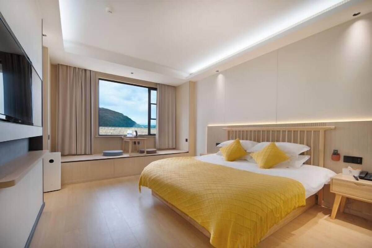 A modern hotel room with a large bed, a bedside table, a desk, and a view of the outdoors.