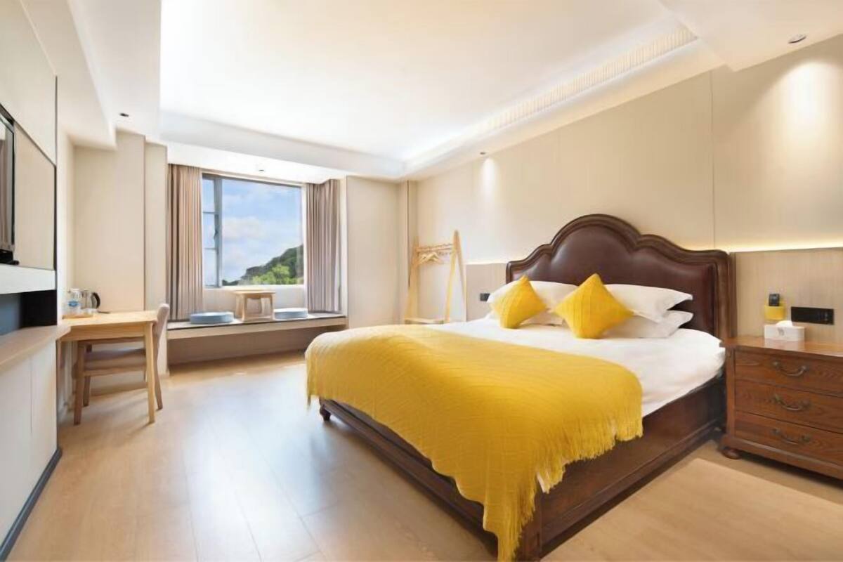 A modern hotel room with a large bed, a wooden nightstand, a desk, and a window with a view of greenery.
