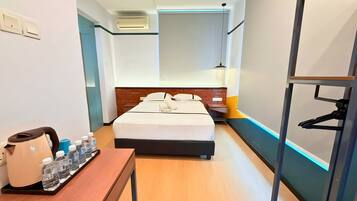 A hotel room with a bed, bedside table with water bottles, a kettle, a hanging light fixture, and a coat rack.