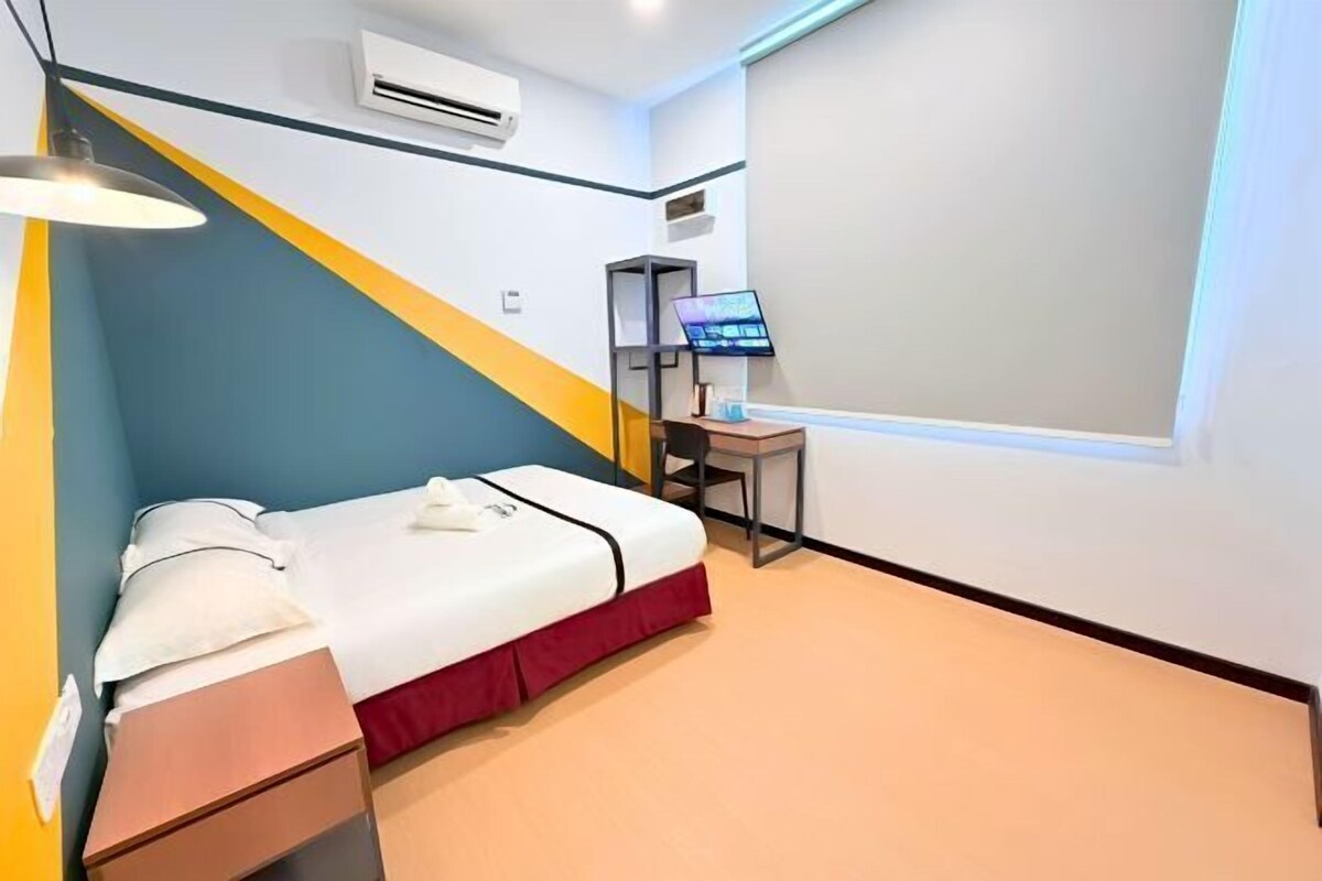 A hotel room with a bed, a desk with a computer, an air conditioner, and a window.