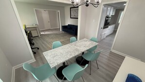 Un comedor moderno con una mesa de mármol y sillas color turquesa, una araña, y una cocina al fondo.