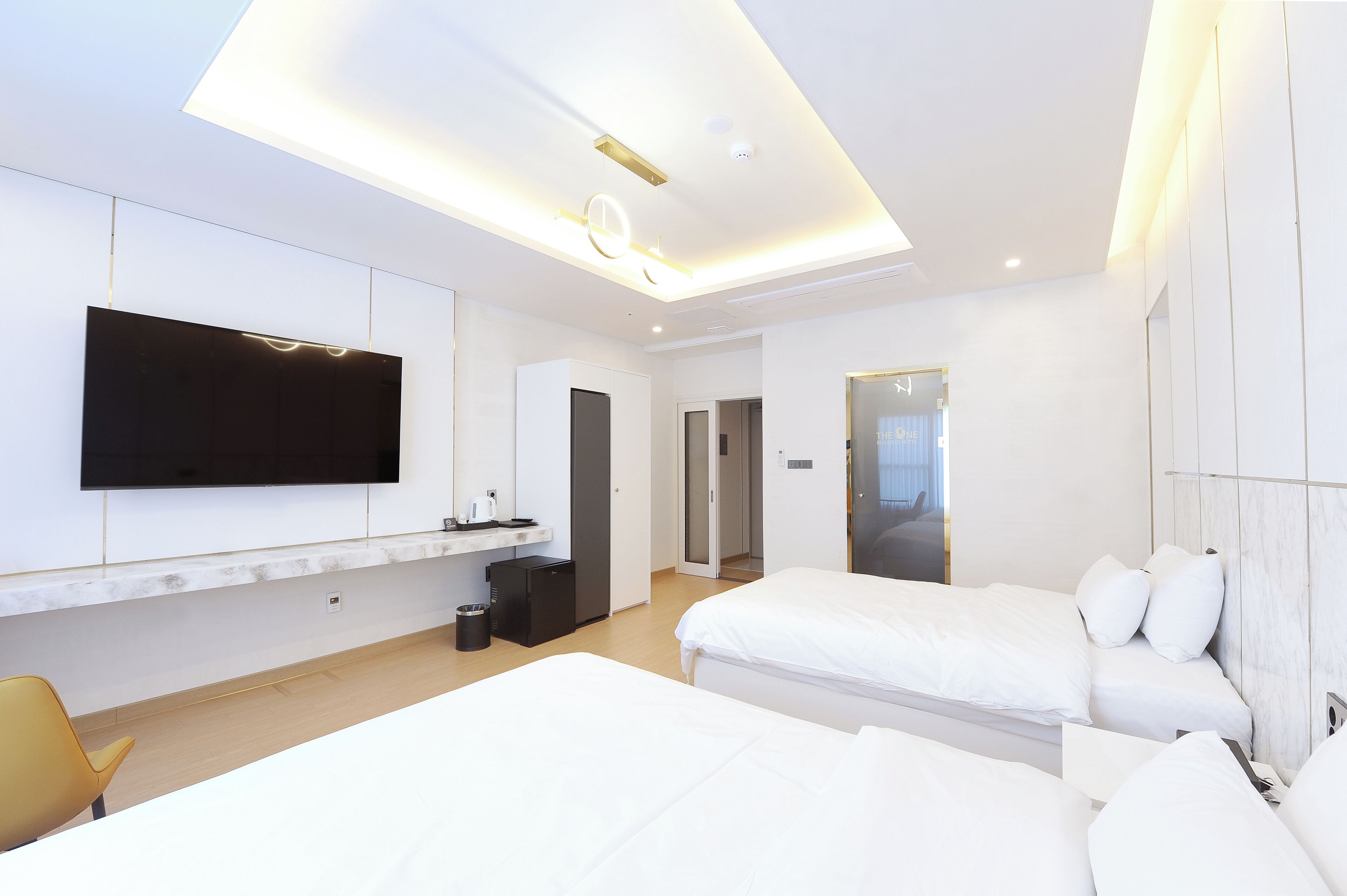A modern hotel room with a large flat-screen TV mounted on the wall, a small kitchenette, two beds, and a separate bathroom.