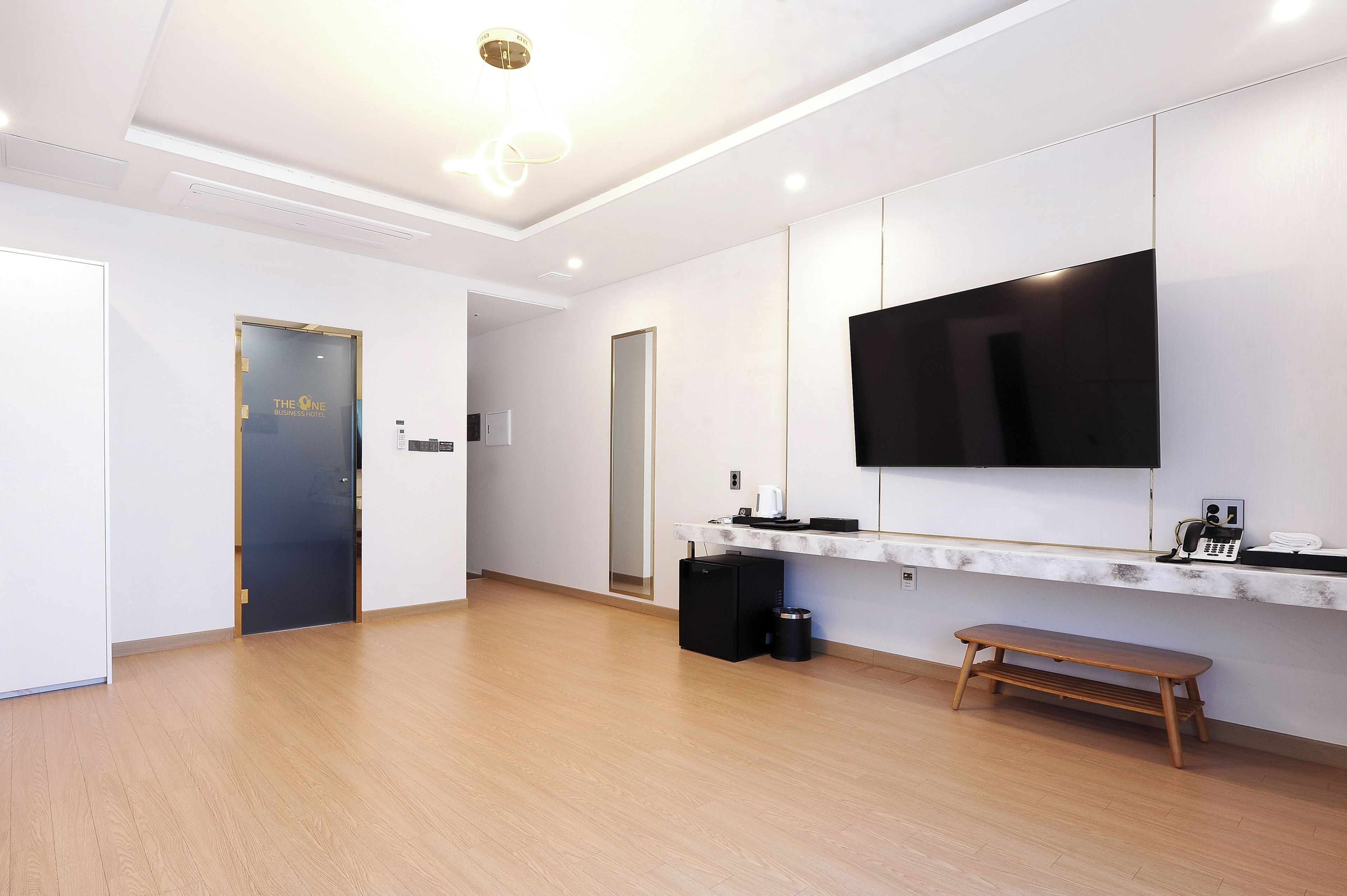 Una habitación de hotel moderna con una gran televisión de pantalla plana montada en la pared, un pequeño escritorio negro con un teléfono y una cafetera, un banco de madera y una puerta con marco dorado.