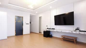 Una habitación de hotel moderna con una gran televisión de pantalla plana montada en la pared, un pequeño escritorio negro con un teléfono y una cafetera, un banco de madera y una puerta con marco dorado.
