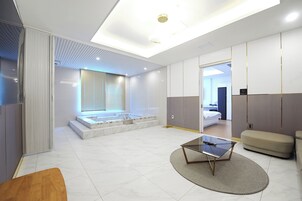 Una habitación de hotel moderna con una cama grande, una zona de estar con un sofá, una mesita de cristal y un baño pequeño con bañera.