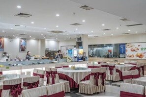 A banquet hall with round tables and chairs, a counter with food display, and a person in the background.