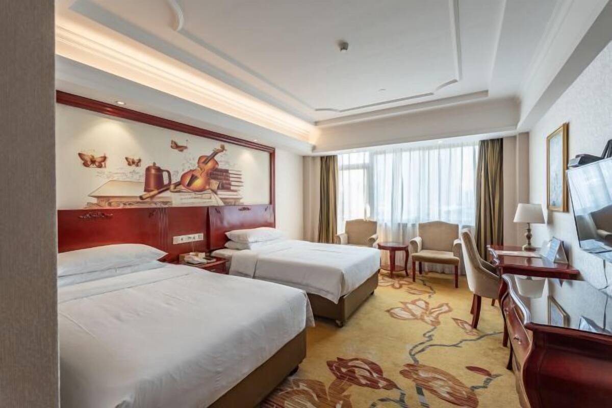A hotel room with two beds, a desk, a chair, a TV, and a painting on the wall.