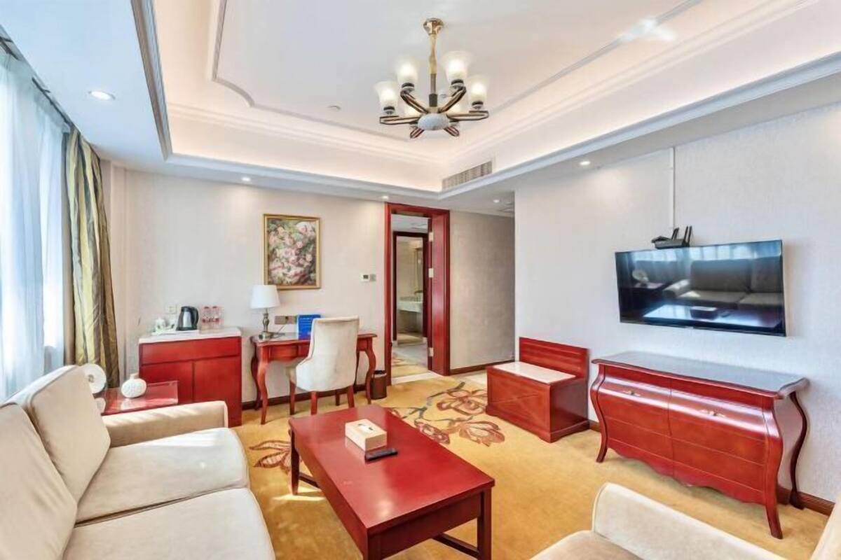 A hotel room with a sofa, a red coffee table, a TV mounted on the wall, and a desk with a chair.
