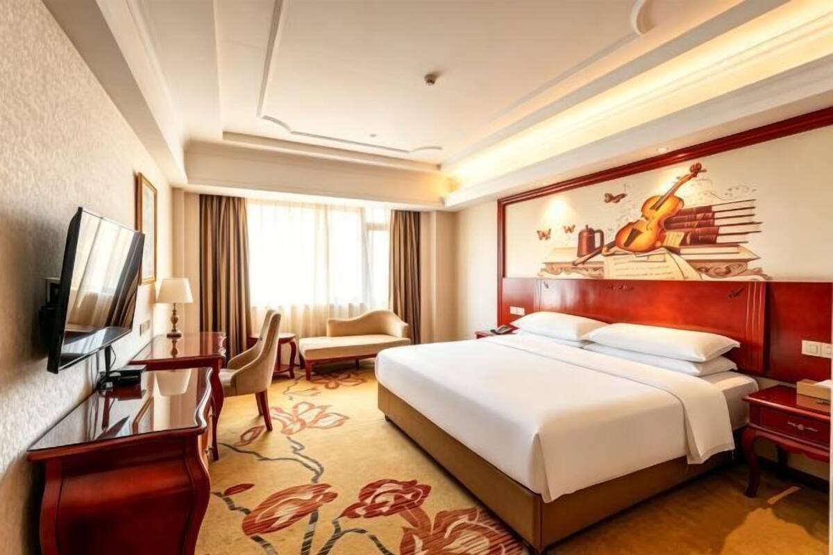 A hotel room with a bed, a desk with a TV, a chair, a sofa, and a wall with a musical instrument and books.