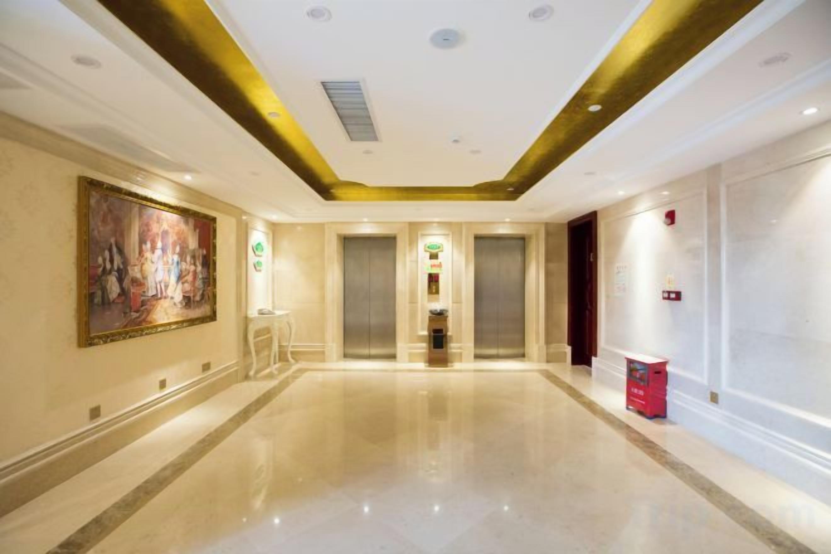 A hotel corridor with a polished floor, a large painting on the left, and two elevators.